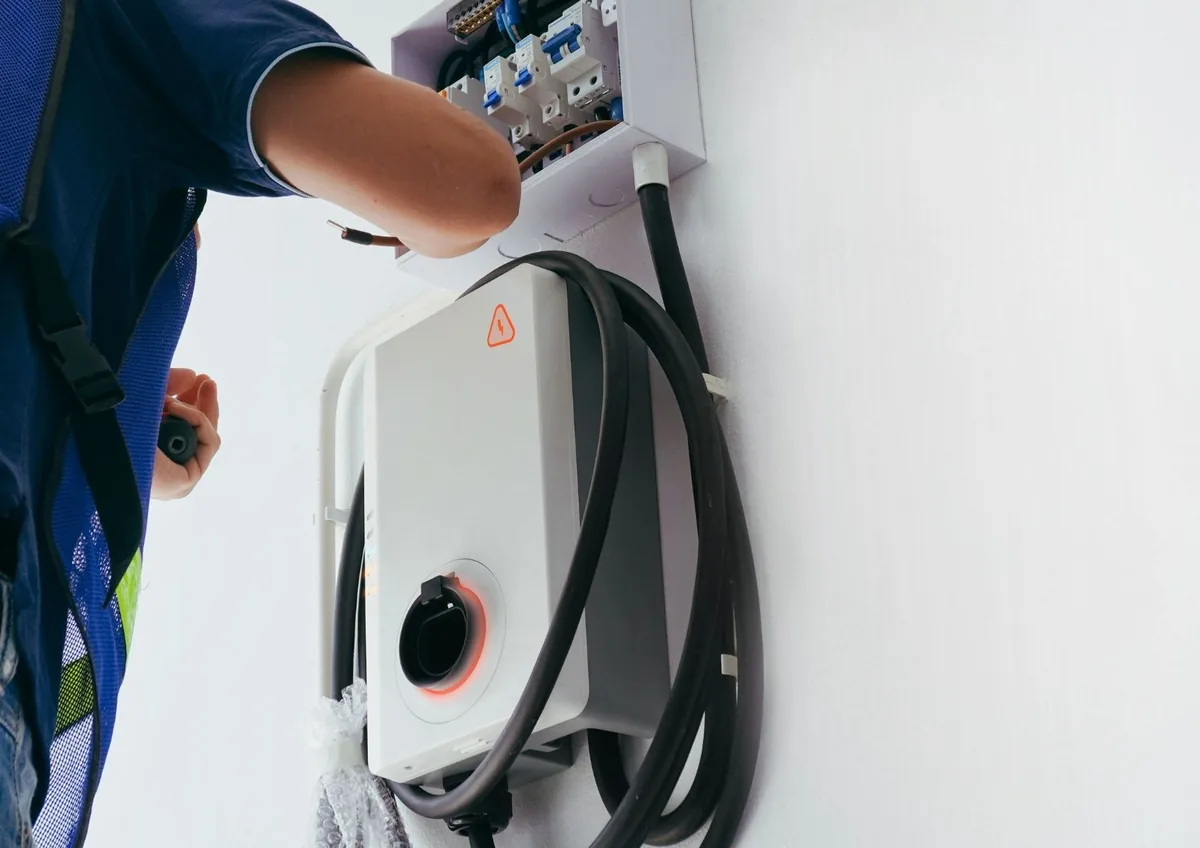 Electrician in a safety vest wiring a small breaker box above a wall-mounted EV charger with its cable coiled